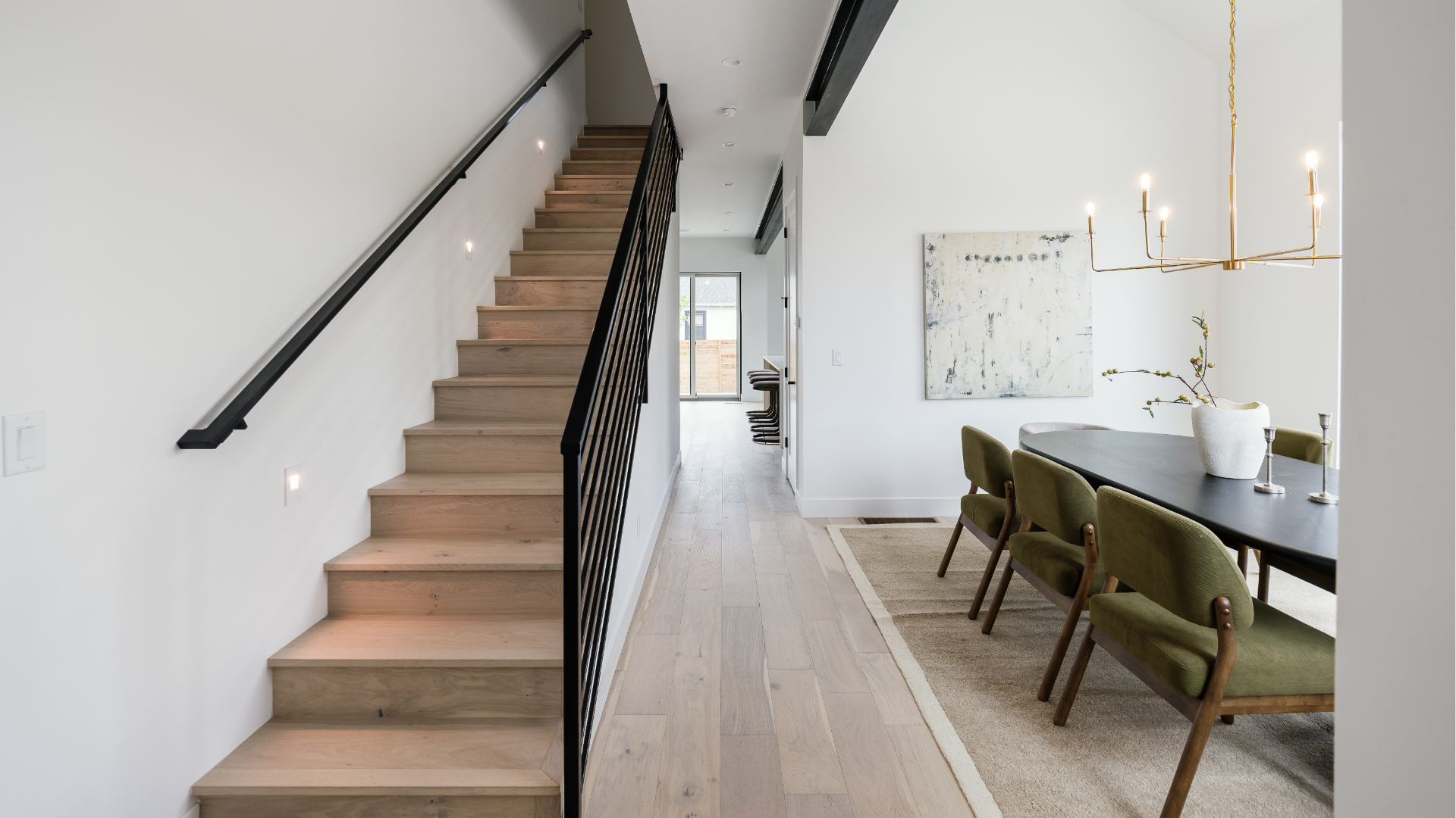 Open oak staircase with iron railing near a dining room with abstract art and brass chandelier